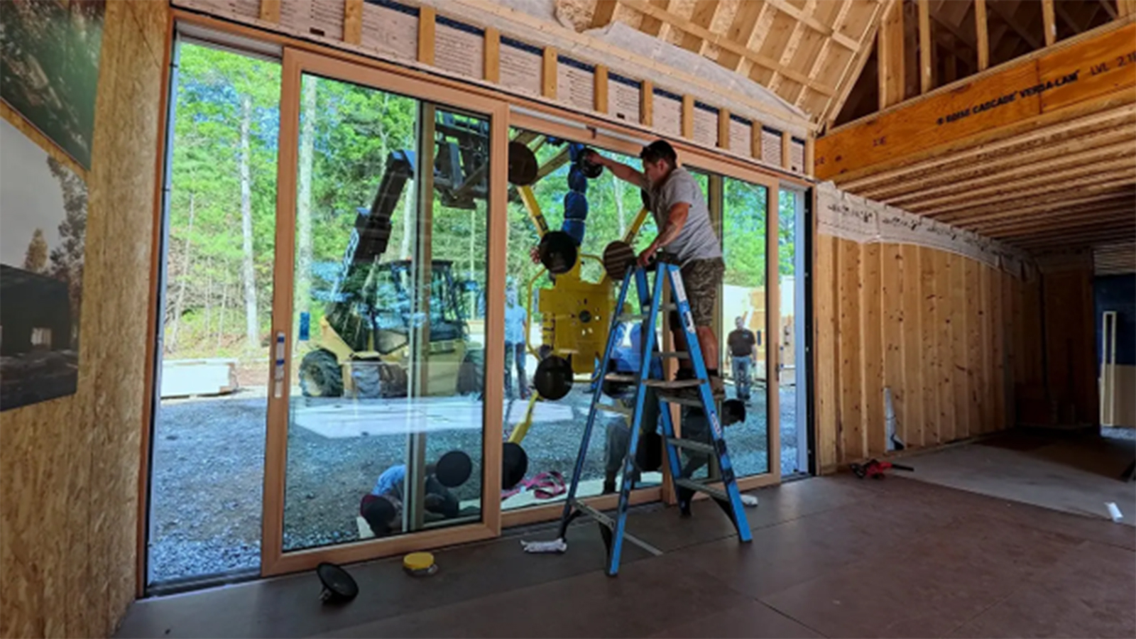 Workers install large glass door panels inside a wood-framed cabin under construction.